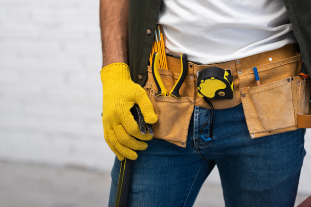 Cropped view of man in glove and tool belt holding hammer at home  - Photo, Image