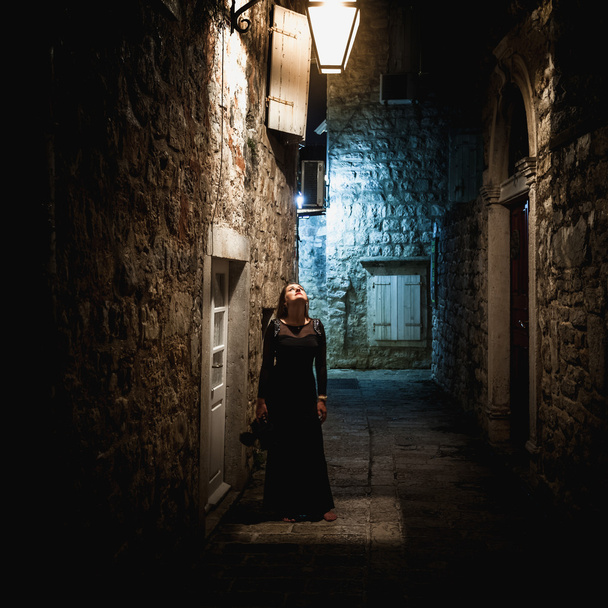 woman in long black dress standing under lantern at old narrow s - Foto, imagen