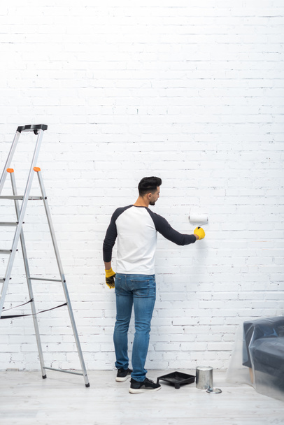 Side view of muslim man in gloves coloring brick wall with roller at home  - Foto, Imagem