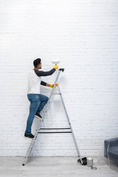 Arabian man coloring brick wall while standing on ladder in living room  - Photo, Image