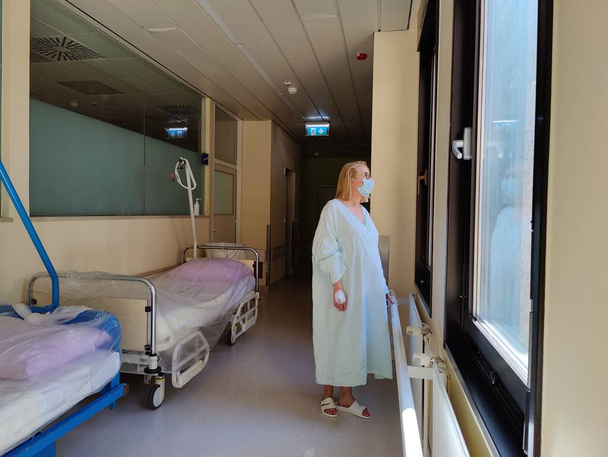 Solitary female patient wearing mask and hospital robe looking out trough big windows of hospital hallway - Foto, Imagen