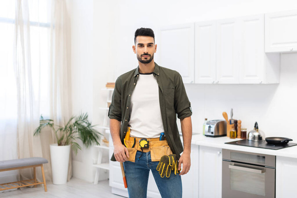 Арабский человек в инструментальном поясе смотрит в камеру на кухне  - Фото, изображение