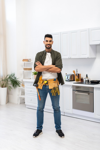 Cheerful muslim man with tool belt crossing arms in kitchen  - Zdjęcie, obraz