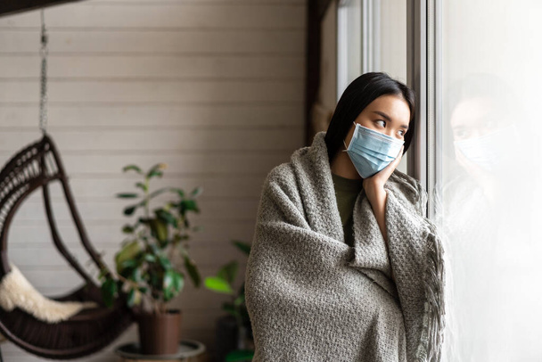 Triste malata ragazza asiatica in quarantena, in piedi vicino alla finestra a casa e guardando fuori, indossando maschera medica da covid. - Foto, immagini