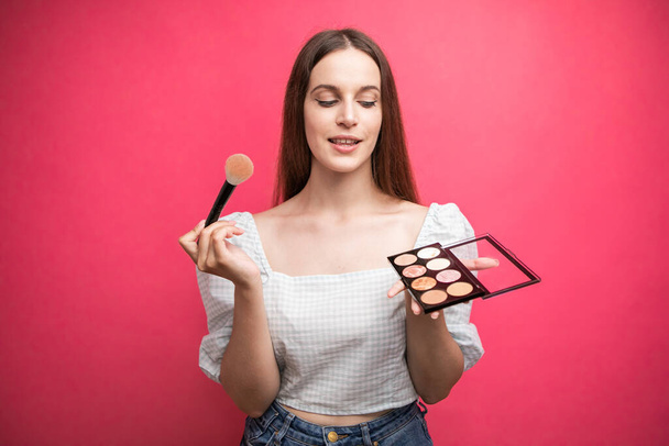 portrait of an attractive lovely young woman standing isolated over pink background, holding makeup blusher brush. High quality photo - Foto, Imagem