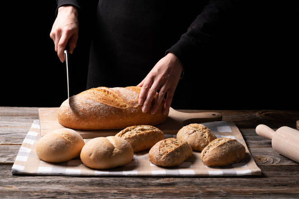 Youn Lady baker cuts her freshly beked bread on the chopping board. Bread and whole grains buns exposed onthe kitchen cloth placed on the wooden surface. - Foto, immagini