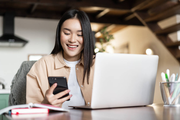 Улыбающаяся азиатка-студентка делает домашнее задание на ноутбуке. Женщина использует мобильный телефон и компьютер для работы из дома. - Фото, изображение