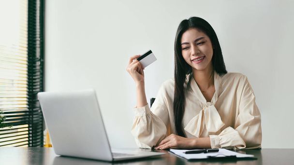 Happy young woman holding credit card and using laptop for making payment online. Online shopping, e-commerce concept. - Fotó, kép