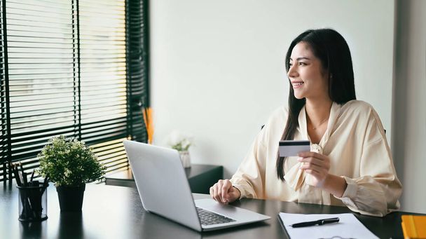 Smiling young woman holding credit card, making payment on internet via laptop computer. Online shopping, e-commerce concept. - 写真・画像