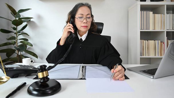 Зріла жінка-адвокат у халаті сидить перед ноутбуком і розмовляє по телефону зі своїми клієнтами для надання юридичної консультації
. - Фото, зображення
