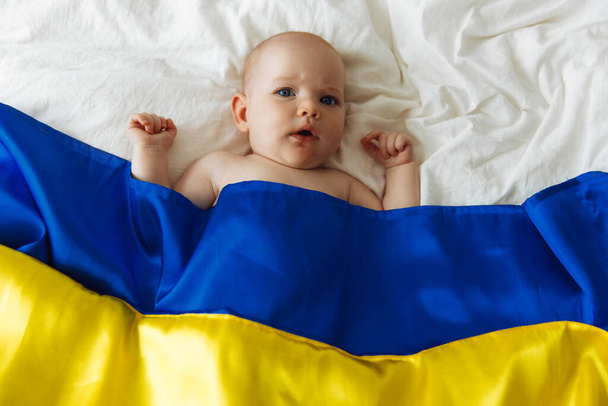 Portrait of a baby wrapped in the national blue and yellow flag of Ukraine, lying on the bed. Baby with Ukraine flag heart. photo with place for text. Children of war. The child is scared. Hope for the world. fear and expectation. children's tears. S - Fotografie, Obrázek