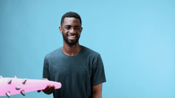 Afrykański Amerykanin bawi się nadmuchiwanym różowym kijem w dłoniach, uderza się w głowę, niedbale tańczy w koszulce na niebiesko. tło w studio. Wysokiej jakości materiał 4k - Materiał filmowy, wideo