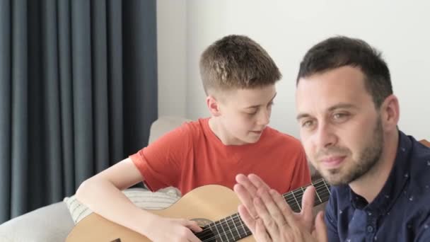 A teenage boy plays the guitar in the room with his dad. Dad teaches his son to play the guitar. Family time together - Imágenes, Vídeo