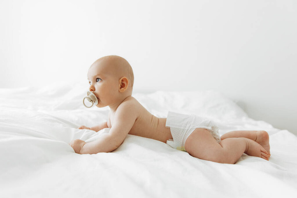 newborn baby sucks on a pacifier and lies on a white bed at home in a white sunny bedroom. Textiles and bed linen for children. Happy healthy baby concept, space for text - Valokuva, kuva