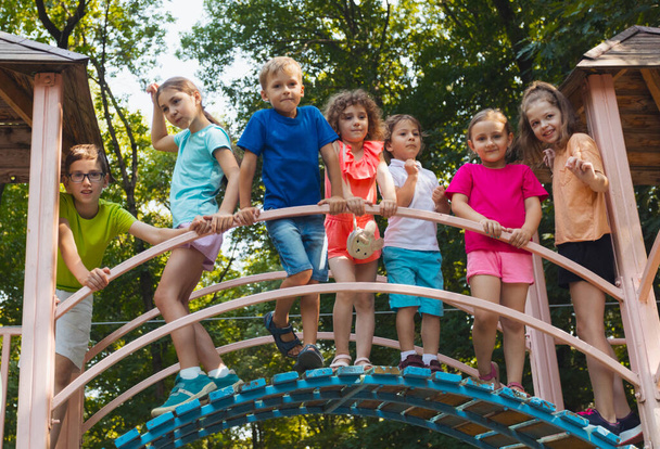The elementary school children stand on a bridge in the playground and hold on to the railing. The kids dressed in colorful clothes are posing for the camera - Zdjęcie, obraz