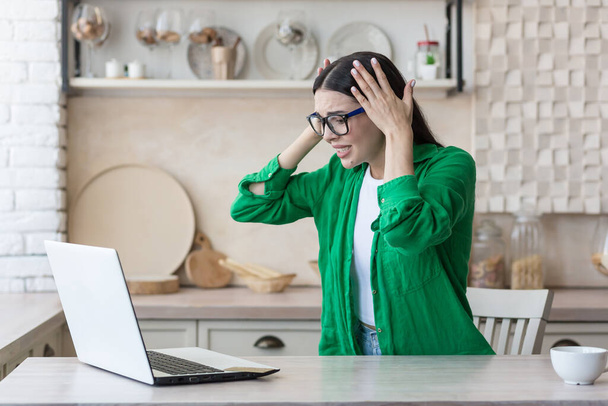 Stressed woman at home working in kitchen using laptop, got bad news online, brunette holding her head sad - Fotoğraf, Görsel