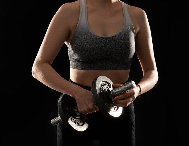 Close up image of slim and sporty woman with steel dumbbell in hand on the black background - Zdjęcie, obraz
