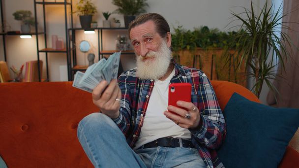 Планирую семейный бюджет. Улыбающийся дедушка считает деньги, использует смартфон, рассчитывает домашние счета дома. Радостный человек доволен доходом и экономит деньги на запланированный отпуск, подарки - Фото, изображение