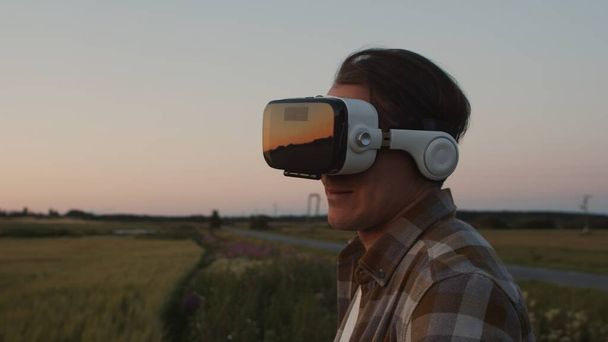 Farmer in virtual reality helmet in front of a sunset agricultural landscape. Man in a countryside field. The concept of country life, food production, farming and technology. - Photo, image