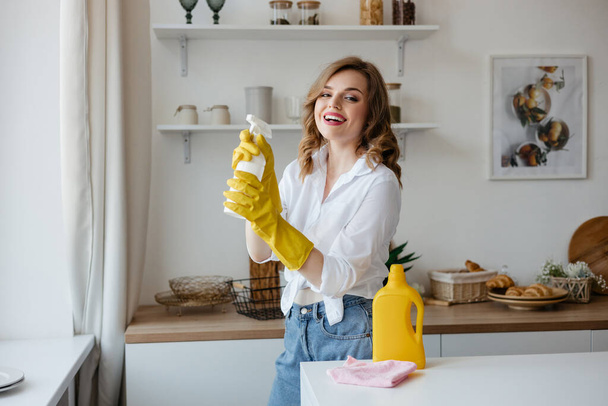 Beautiful girl in yellow gloves holding a cleaning spray in the kitchen. High quality photo - Foto, Bild