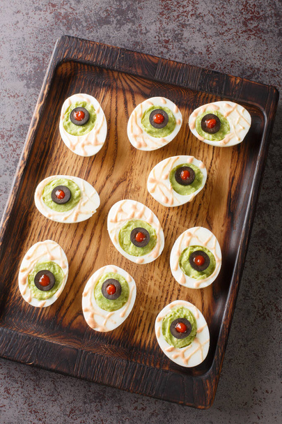 Guacamoldy Eyeballs Eggs stuffed with avocado cream in a wooden tray on the table. Vertical top view from abov - Photo, image