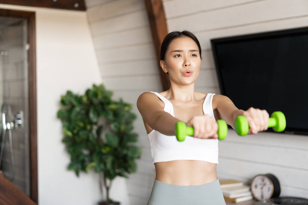 Fitness aziatische meisje doet workout thuis in de woonkamer, tillen halters, het dragen van activewear voor sport activiteiten. - Foto, afbeelding