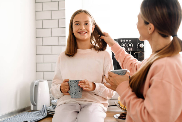 White mother and daughter talking and drinking tea in kitchen at home - Photo, Image