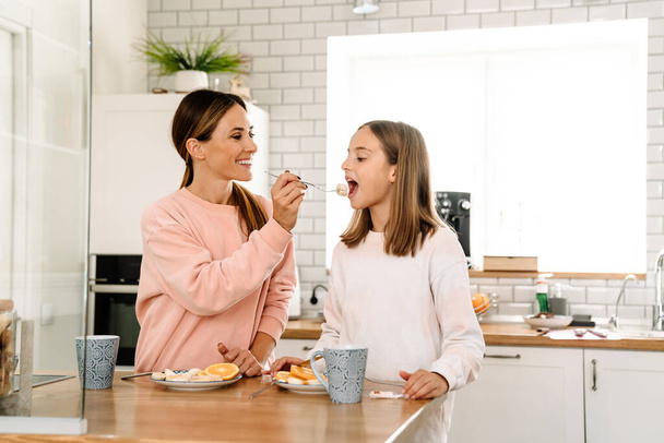 White mother and daughter smiling while having breakfast in kitchen at home - 写真・画像