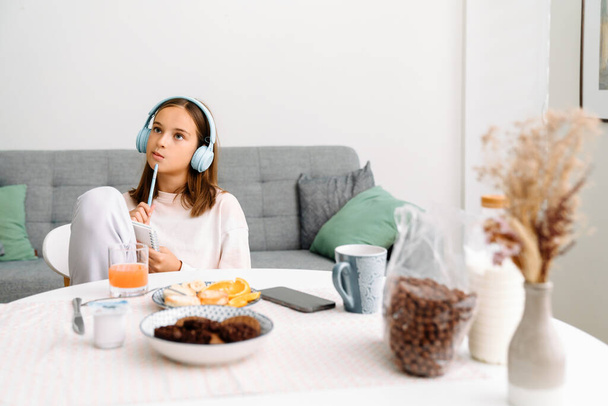 White girl in headphones doing homework while having breakfast at home - Фото, изображение