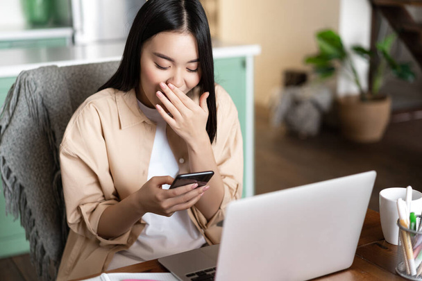 Boldog ázsiai lány ül laptoppal otthon, nézi a mobiltelefon és nevet, munkahelyi. - Fotó, kép
