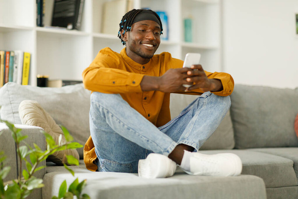 Happy african man using phone while sitting on sofa at his home. Concept of young people working on mobile devices. High quality photo - Foto, Imagem
