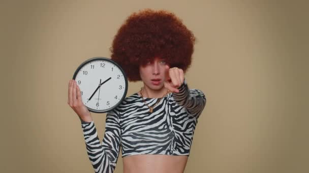 It is your time. Pretty young woman with brown lush wig showing time on wall office clock, ok, thumb up, approve, pointing finger at camera. Adult stylish female girl on beige studio background indoor - Imágenes, Vídeo