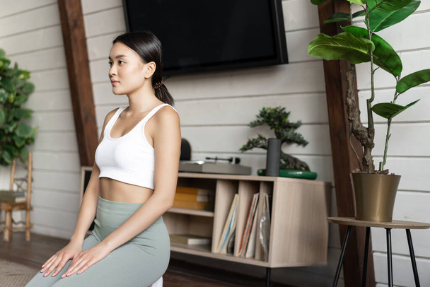 Bild einer asiatischen Frau, die sich während der Morgenmeditation konzentriert, Yoga zu Hause praktiziert, im Wohnzimmer sitzt, Zen-Pose einnimmt. - Foto, Bild