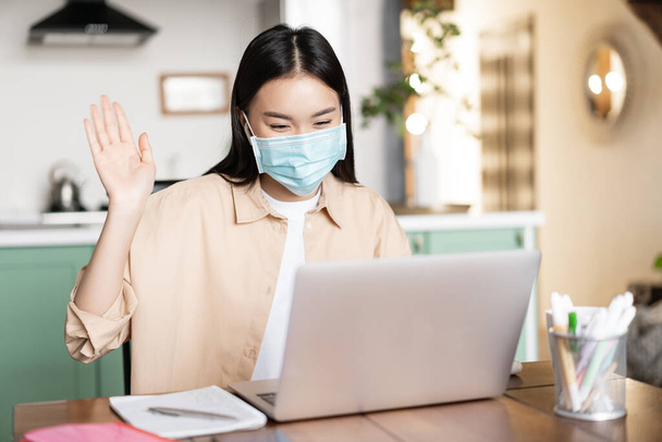 Jonge Aziatische vrouw die op afstand werkt en sociale afstand neemt. Meisje in medisch masker zwaaien op video conferentie op laptop. - Foto, afbeelding