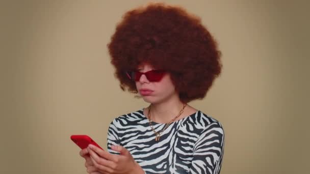 Sad young woman with brown lush wig use smartphone typing browsing, loses becoming surprised sudden lottery results, bad fortune, loss. Adult stylish girl isolated on beige studio background indoors - Πλάνα, βίντεο