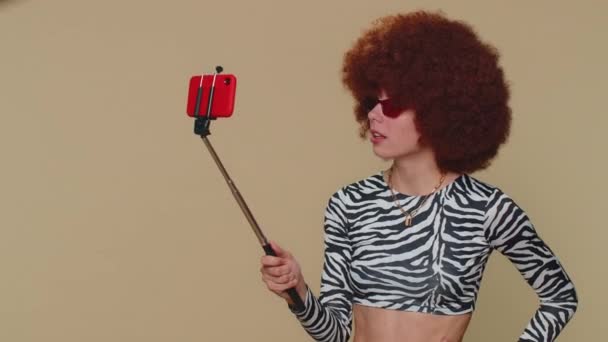 Wow reaction. Impressed pretty young woman with brown lush wig blogger taking selfie on mobile phone selfie stick, communicating video call online. Adult stylish girl on beige studio background indoor - Video