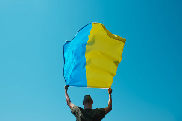чоловік, побачений ззаду, хвилює український прапор над головою на небі в сонячний день
 - Фото, зображення