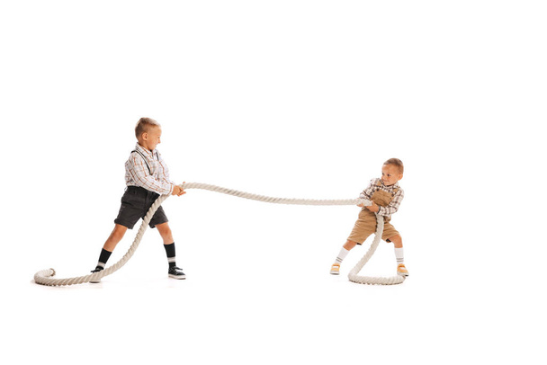 Portrait of playful children, two stylish boys in vintage clothes pulling the rope isolated over white studio background. Concept of childhood, friendship, fun, lifestyle, fashion. Retro style - Фото, изображение