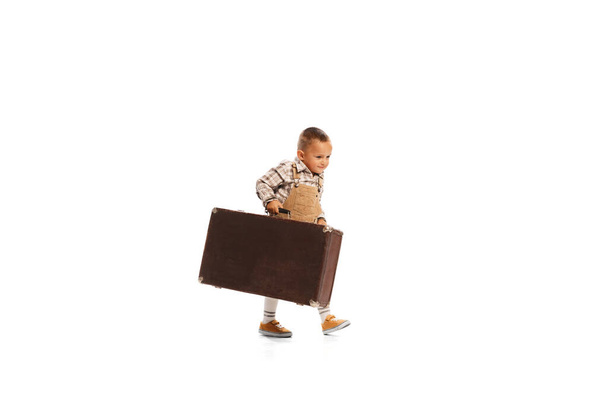 Portrait of little boy, child carrying giant vintage suitcase isolated over white studio background. Travelling . Concept of childhood, friendship, fun, lifestyle, fashion. Retro style - Photo, Image
