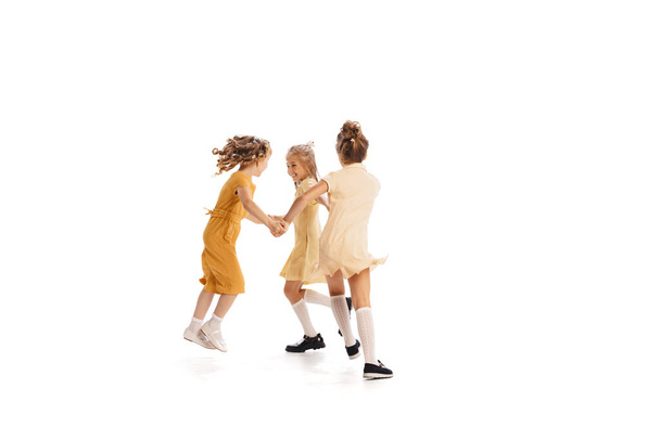 Портрет трьох маленьких дівчат в жовтих стильних сукнях, які грають разом ізольовано над білим фоном студії. Кумедний спосіб життя. Концепція дитинства, дружби, веселощів, способу життя, моди. Стиль ретро - Фото, зображення