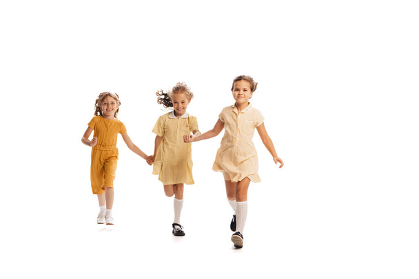 Portrait of three little girls in yellow stylish dresses playing together isolated over white background. Cheerfully running. Concept of childhood, friendship, fun, lifestyle, fashion. Retro style - Foto, Imagem