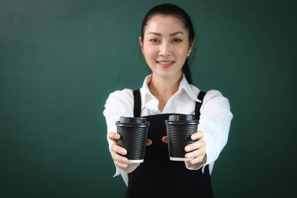 Black paper disposable coffee cups on hand of beautiful Asian female barista wearing apron, female giving two coffee cups to camera on green background, smiling pretty waitress staff working at cafe. - Valokuva, kuva