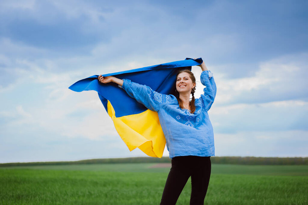Happy free ukrainian woman with national flag on dramatic sky background. Portrait of lady in blue embroidery vyshyvanka shirt. Ukraine, independence, patriot symbol. High quality photo - Foto, afbeelding