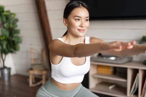 Jeune femme de fitness asiatique avec un corps en bonne santé, faire des squats, entraînement matinal, porter des vêtements de sport, debout à la maison dans le salon et ayant une session d'entraînement. - Photo, image