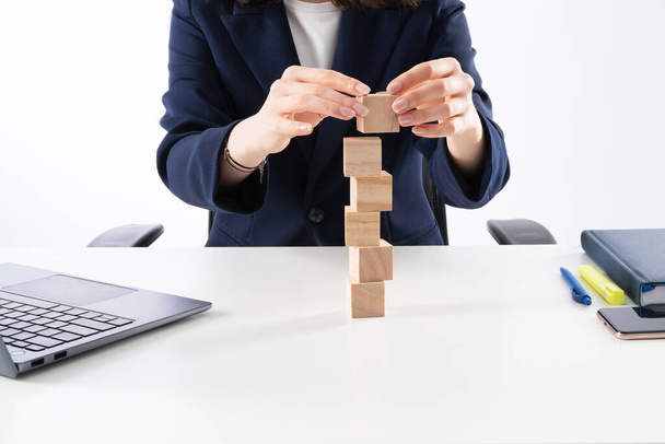 A close up image of bussines woman woman stacking wooden blocks in her work enviroment. Office interior laptop, glass of water and notebook on white desk. - Photo, image
