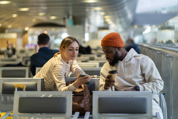 Beautiful European woman asks handsome African American man. International couple reading information online on smartphone, expresses opinion on issue, help connect wifi or booking apartment online. - Foto, Bild