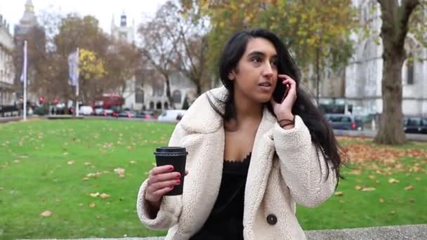 Hermosa chica india hace una llamada telefónica mientras está en el centro de Londres y tiene una taza de café en la mano - Metraje, vídeo