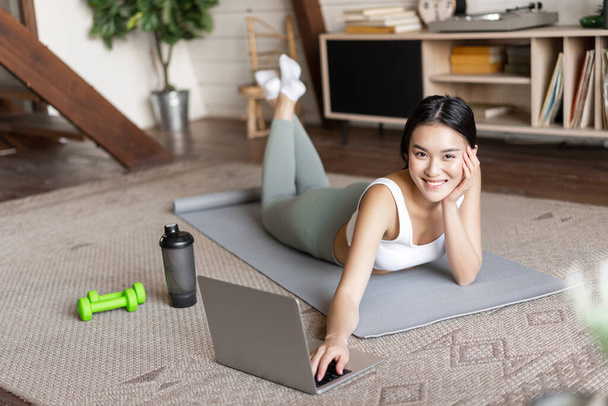 Aasialainen fitness-tyttö ottaa online-kuntokurssin, katselee urheiluvideota, treenaa kotona lattialla matto kanssa dumbells ja proteiini ravistin pullo, hymyillen kameralle. - Valokuva, kuva