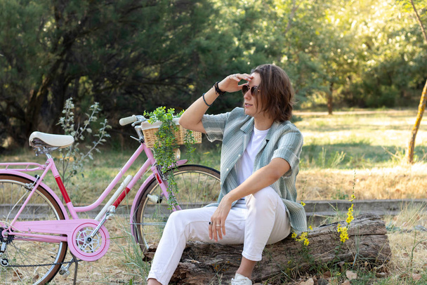 Young man looking far with hand on face sitting in the park and his pink vintage bike behind with green background - Foto, Imagem