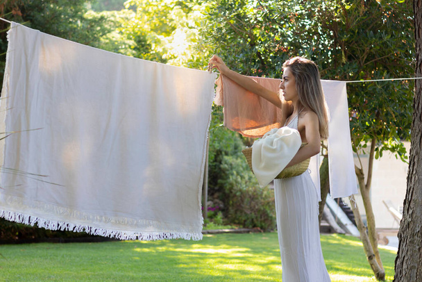 Genç ve güzel bir kadın beyaz bir elbiseyle bahçedeki çamaşırları asıyor ve yazın etrafı doğa ile çevrili. - Fotoğraf, Görsel